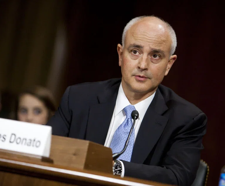 TechDogs-"Judge James Dunato in a suit speaking into a microphone during a formal hearing, seated at a desk with his nameplate visible."